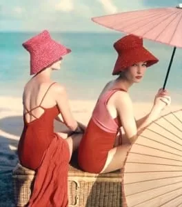 Editorial vintage: dos modelos con sombreros de rafia rojos y trajes de baño rojo y rosa bajo una sombrilla en la playa